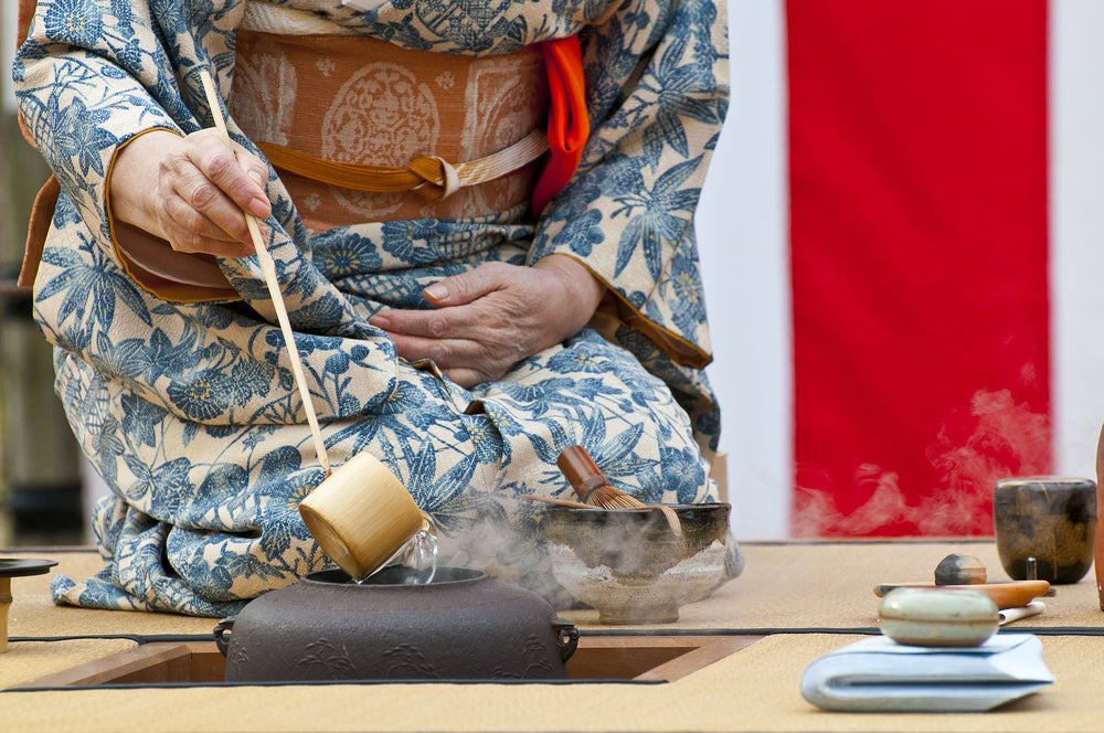 An image of traditional Japanese tea brewing. An individual is sitting on a .mat, brewing tea with Japanese tea equipment