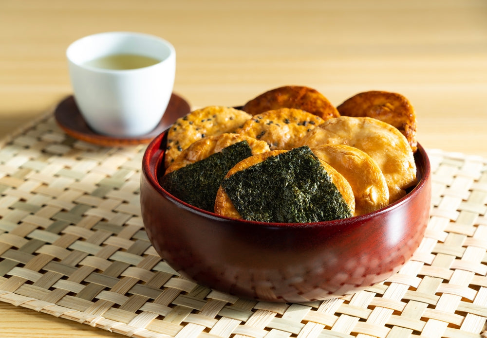 A bowl of senbei rice crackers beside a cup of tea on a wooden placemat.